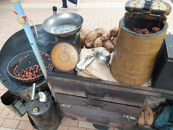 A street food cart with roasted chestnuts displayed in a pan over charcoals. A traditional weighing scale and a cylindrical wooden container filled with sweet potatoes are also visible. The equipment is rustic and appears used, placed on a paved surface.