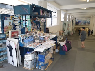 A small kiosk located in a covered walkway selling various magazines, newspapers, bottled water, and other small items. The kiosk is cluttered with publications such as fashion magazines and newspapers stacked on a table and in shelves. Some graffiti is visible on the kiosk. People are walking nearby in a casual setting.