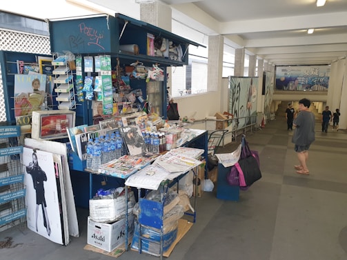 A small kiosk located in a covered walkway selling various magazines, newspapers, bottled water, and other small items. The kiosk is cluttered with publications such as fashion magazines and newspapers stacked on a table and in shelves. Some graffiti is visible on the kiosk. People are walking nearby in a casual setting.