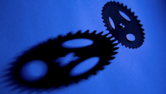 Two interconnected gears are set against a blue background, casting dark shadows that feature distinct shapes and cutouts.