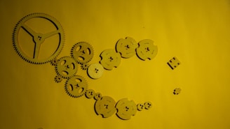 A collection of interlocking wooden gears arranged on a yellow background. The gears vary in size and are positioned to form a sequence that appears as if they could rotate together.