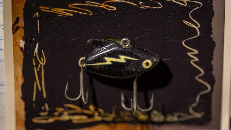A vintage fishing lure with a black body, featuring a yellow zigzag pattern and a circular eye design. The lure is equipped with two metal hooks and is displayed against a dark backdrop adorned with abstract yellow scribbles and designs.