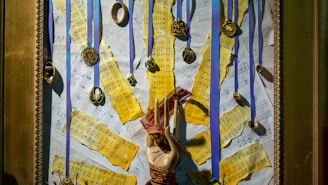 A framed collage featuring yellowed sheet music pieces torn into strips and arranged over a background of more music sheets. Multiple medals and circular ornaments hang from blue ribbons attached to the top of the frame. In the center, a hand sculpture wrapped with red thread stands out against the background.