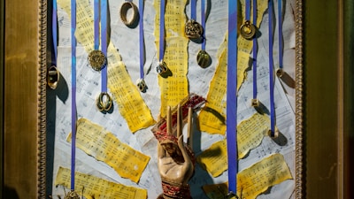 A framed collage featuring yellowed sheet music pieces torn into strips and arranged over a background of more music sheets. Multiple medals and circular ornaments hang from blue ribbons attached to the top of the frame. In the center, a hand sculpture wrapped with red thread stands out against the background.