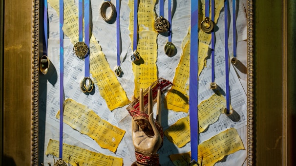 A framed collage featuring yellowed sheet music pieces torn into strips and arranged over a background of more music sheets. Multiple medals and circular ornaments hang from blue ribbons attached to the top of the frame. In the center, a hand sculpture wrapped with red thread stands out against the background.