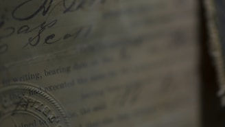 A close-up view of an old, textured document with handwritten and printed text. The text is partially visible and includes a seal or stamp embossed on the paper. The overall appearance is aged, with a sepia tone, contributing to a historical feel.