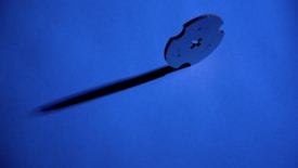 A metal blade stands upright on a blue background, casting a long shadow. The blade has a cross-shaped cutout in the center and curved edges.