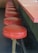 A series of red, round bar stools aligned along a countertop, suggesting a retro or vintage diner style. The stools have metal supports and are positioned on a dark floor, paired with a matching countertop that has a pinkish hue.