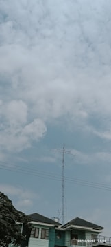 A tall antenna stands atop a building with a clear blue sky and scattered clouds overhead. The structure appears to be used for broadcasting or communication, and the building roof is partially visible with a nearby leafy tree.