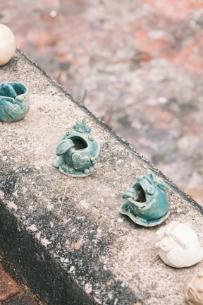 Small ceramic sculptures are displayed on a rough concrete surface. The sculptures are primarily blue-green and appear to depict playful and whimsical creatures or animals, some with detailed patterns or designs. The background is blurred and features earthy tones, creating a rustic outdoor setting.