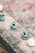 Small ceramic sculptures are displayed on a rough concrete surface. The sculptures are primarily blue-green and appear to depict playful and whimsical creatures or animals, some with detailed patterns or designs. The background is blurred and features earthy tones, creating a rustic outdoor setting.