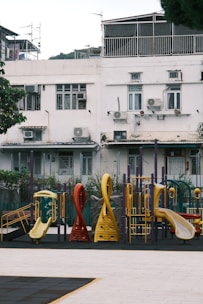A vibrant children's playground situated in front of an old, white residential building. The playground features colorful slides and climbing structures in shades of yellow, red, and green. The building behind has multiple windows, air conditioning units, and visible signs of aging with peeling paint and some structural wear.