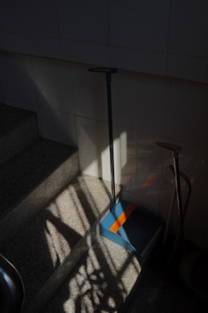 A dimly lit stairway area with sunlight partially illuminating the steps. A dustpan with a long handle is resting against the tiled wall on one of the steps. Shadows of window frames or bars create patterns on the steps and wall. There are also some cleaning tools like a broom placed leaning against the wall.