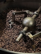 A large quantity of roasted coffee beans surrounds a metal mechanism inside a coffee roaster. The device appears industrial with several bolts and a rotating arm, indicative of the process of roasting coffee beans.