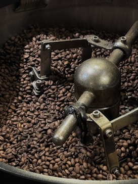 A large quantity of roasted coffee beans surrounds a metal mechanism inside a coffee roaster. The device appears industrial with several bolts and a rotating arm, indicative of the process of roasting coffee beans.