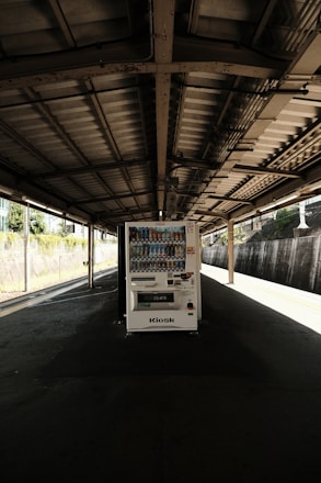 A solitary vending machine stands under a dimly lit, industrial-style train platform. The scene is quiet and empty, with shadows cast by the overhead metal roof. Sunlight filters in from the sides, partially illuminating the vending machine and concrete ground.