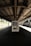A solitary vending machine stands under a dimly lit, industrial-style train platform. The scene is quiet and empty, with shadows cast by the overhead metal roof. Sunlight filters in from the sides, partially illuminating the vending machine and concrete ground.