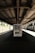 A solitary vending machine stands under a dimly lit, industrial-style train platform. The scene is quiet and empty, with shadows cast by the overhead metal roof. Sunlight filters in from the sides, partially illuminating the vending machine and concrete ground.