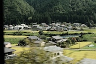 A small rural village nestled among lush green fields and forests. Traditional houses with sloped roofs are scattered throughout the landscape, with a backdrop of dense trees on a hillside.