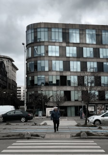 An individual wearing a jacket and cap, walking on a crosswalk in front of a modern multi-story office building. The building features large glass windows and has a curved architectural design. The sky is overcast, and there are cars parked along both sides of the street. Leafless trees line the sidewalk, and a few pedestrians are visible in the background.