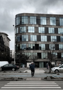 An individual wearing a jacket and cap, walking on a crosswalk in front of a modern multi-story office building. The building features large glass windows and has a curved architectural design. The sky is overcast, and there are cars parked along both sides of the street. Leafless trees line the sidewalk, and a few pedestrians are visible in the background.