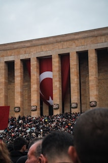 A large crowd gathers outside a monumental building with tall columns and a large Turkish flag hanging between them. The setting suggests a public event or gathering. The crowd, predominantly made up of people dressed in dark colors, fills the foreground under an overcast sky.