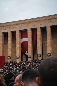 A large crowd gathers outside a monumental building with tall columns and a large Turkish flag hanging between them. The setting suggests a public event or gathering. The crowd, predominantly made up of people dressed in dark colors, fills the foreground under an overcast sky.