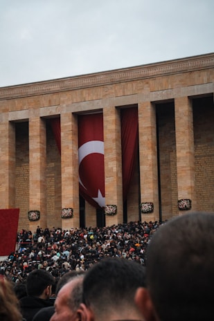 A large crowd gathers outside a monumental building with tall columns and a large Turkish flag hanging between them. The setting suggests a public event or gathering. The crowd, predominantly made up of people dressed in dark colors, fills the foreground under an overcast sky.