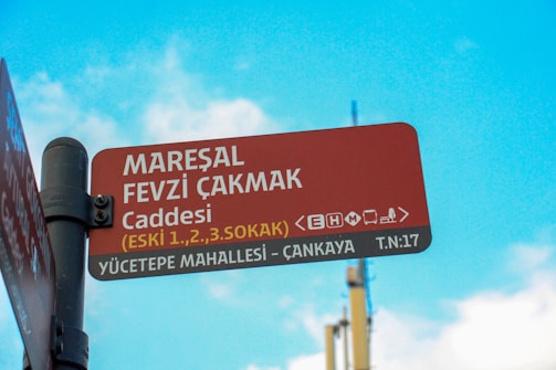 A street sign is mounted on a metal pole against a bright turquoise sky with scattered clouds. The sign is red with white and yellow text, providing street names and directions. It includes the name 'Mareşal Fevzi Çakmak Caddesi' and additional information such as '<eHm>' and 'Yücetepe Mahallesi – Çankaya T.N: 17'.