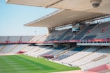 A large, empty sports stadium features rows of gray and white seats, with a field of green grass in the center. The stadium is covered partially by a modern roof, and banners and advertisements are visible around the seating areas.