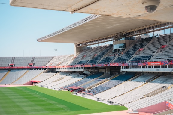 A large, empty sports stadium features rows of gray and white seats, with a field of green grass in the center. The stadium is covered partially by a modern roof, and banners and advertisements are visible around the seating areas.