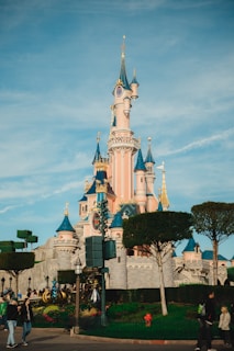 A fairytale-style castle with pink and blue turrets surrounded by neatly trimmed trees and lush gardens. Visitors are walking leisurely around the area, enjoying the sunny day.