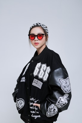 A person wearing a black varsity jacket with various white embroidered designs, including numbers, a cat, and text. They have on red-tinted sunglasses and a black-and-white checkered headband.