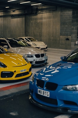 A collection of luxury cars is parked in an indoor garage with a concrete wall. The cars include a bright yellow sports car, a blue vehicle with a distinctive front grille, and a couple of white cars, all showcasing sleek and modern designs. The area is lit by fluorescent lights, casting reflections on the car surfaces.