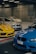 A collection of luxury cars is parked in an indoor garage with a concrete wall. The cars include a bright yellow sports car, a blue vehicle with a distinctive front grille, and a couple of white cars, all showcasing sleek and modern designs. The area is lit by fluorescent lights, casting reflections on the car surfaces.