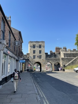 A historic stone gateway with three arches stands prominently in an urban setting, bordered by traditional brick buildings. A person with a backpack walks along the sidewalk under a clear blue sky.