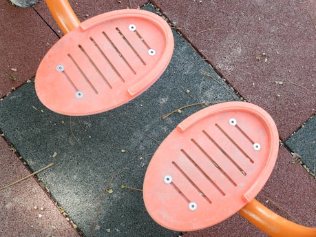 Two orange, oval-shaped footrests with slits are attached to metal bars, set against a background of interlocking rubber tiles in shades of red and gray. The tiles have a textured surface with small debris like twigs scattered across.