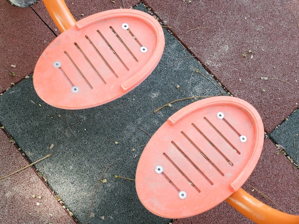 Two orange, oval-shaped footrests with slits are attached to metal bars, set against a background of interlocking rubber tiles in shades of red and gray. The tiles have a textured surface with small debris like twigs scattered across.