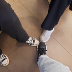 The image shows three people standing together, each displaying one foot in the center. The footwear includes a pair of black Converse sneakers, a pair of gray slip-on sneakers, and a pair of black athletic shoes. The flooring is composed of large, square, brown tiles.