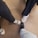 The image shows three people standing together, each displaying one foot in the center. The footwear includes a pair of black Converse sneakers, a pair of gray slip-on sneakers, and a pair of black athletic shoes. The flooring is composed of large, square, brown tiles.