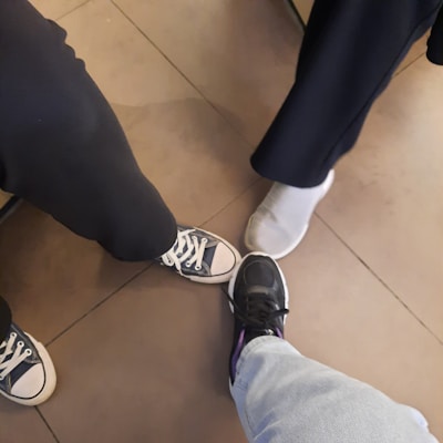 The image shows three people standing together, each displaying one foot in the center. The footwear includes a pair of black Converse sneakers, a pair of gray slip-on sneakers, and a pair of black athletic shoes. The flooring is composed of large, square, brown tiles.