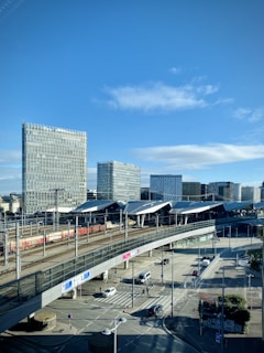 A modern urban setting featuring multiple high-rise buildings with glass facades. A railway station with trains and tracks is prominent in the foreground, while a roadway with vehicles and pedestrians is visible below a bridge. The sky is clear with a few scattered clouds.