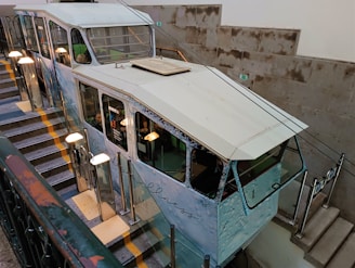 A funicular railway car is positioned on a stairway track inside a building. The car has a vintage design with large windows and appears to be decorated with images or advertising on the sides. The stairway is lined with handrails and poles with globe-shaped lights. The walls are made of concrete blocks, adding an industrial look to the environment.