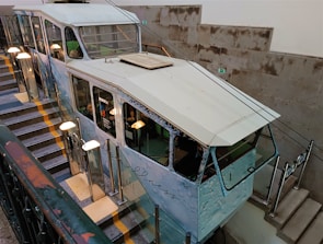 A funicular railway car is positioned on a stairway track inside a building. The car has a vintage design with large windows and appears to be decorated with images or advertising on the sides. The stairway is lined with handrails and poles with globe-shaped lights. The walls are made of concrete blocks, adding an industrial look to the environment.