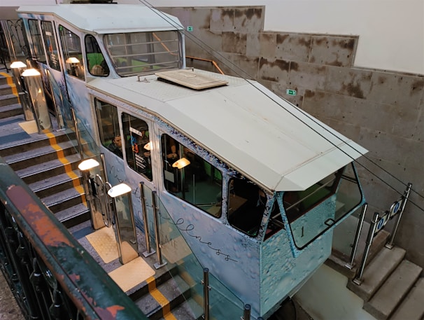 A funicular railway car is positioned on a stairway track inside a building. The car has a vintage design with large windows and appears to be decorated with images or advertising on the sides. The stairway is lined with handrails and poles with globe-shaped lights. The walls are made of concrete blocks, adding an industrial look to the environment.