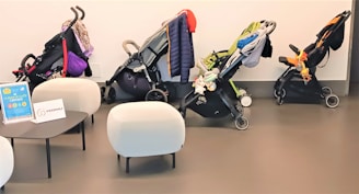 The image depicts a row of three strollers neatly arranged against a light-colored wall. The strollers are equipped with various accessories, such as jackets and toys. In the foreground, there are two modern, white, rounded chairs and a black, low table. Two signs with text are placed on the table.