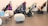 The image depicts a row of three strollers neatly arranged against a light-colored wall. The strollers are equipped with various accessories, such as jackets and toys. In the foreground, there are two modern, white, rounded chairs and a black, low table. Two signs with text are placed on the table.