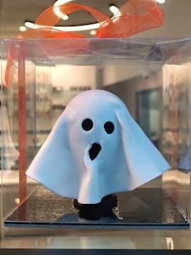 A small ghost figurine is encased in a transparent plastic box, topped with an orange ribbon. The ghost is made of white material and has hollow black eyes and a mouth, giving it a spooky appearance. The background is blurred, indicating an indoor setting with soft lighting.