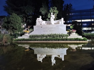 A white marble monument featuring statues, surrounded by lush greenery and flowers, is reflected in a calm pond at night. The scene is set against a backdrop of trees and a modern building.