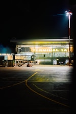 A well-lit airport terminal identified as 'Terminal 1A' with a glass facade. The area in front is an empty tarmac with visible runway markings. A tall light pole illuminates the scene, suggesting night time.